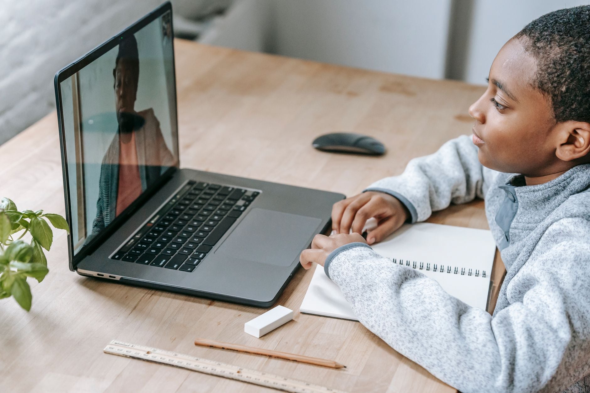 Young student in gray hoodie looking at laptop screen during online lesson at wooden desk with notebook