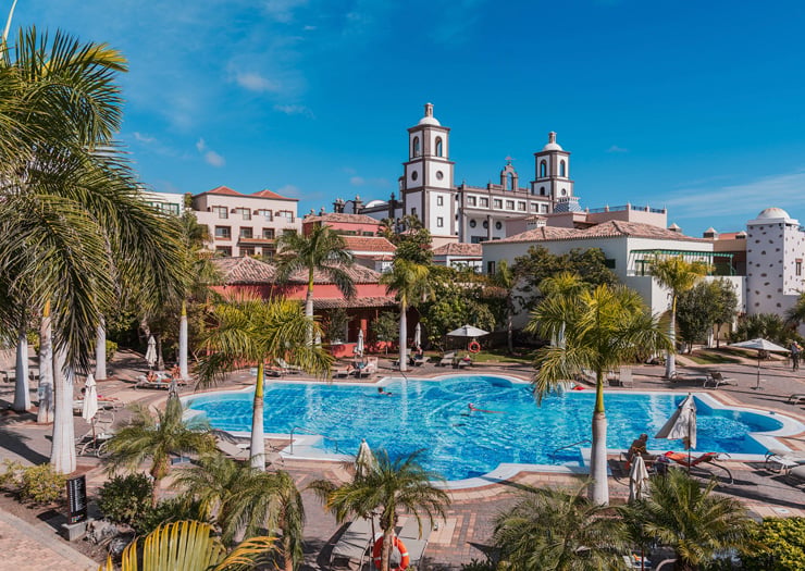 Resort con piscina grande, palmeras y iglesia colonial de dos torres al fondo bajo cielo azul