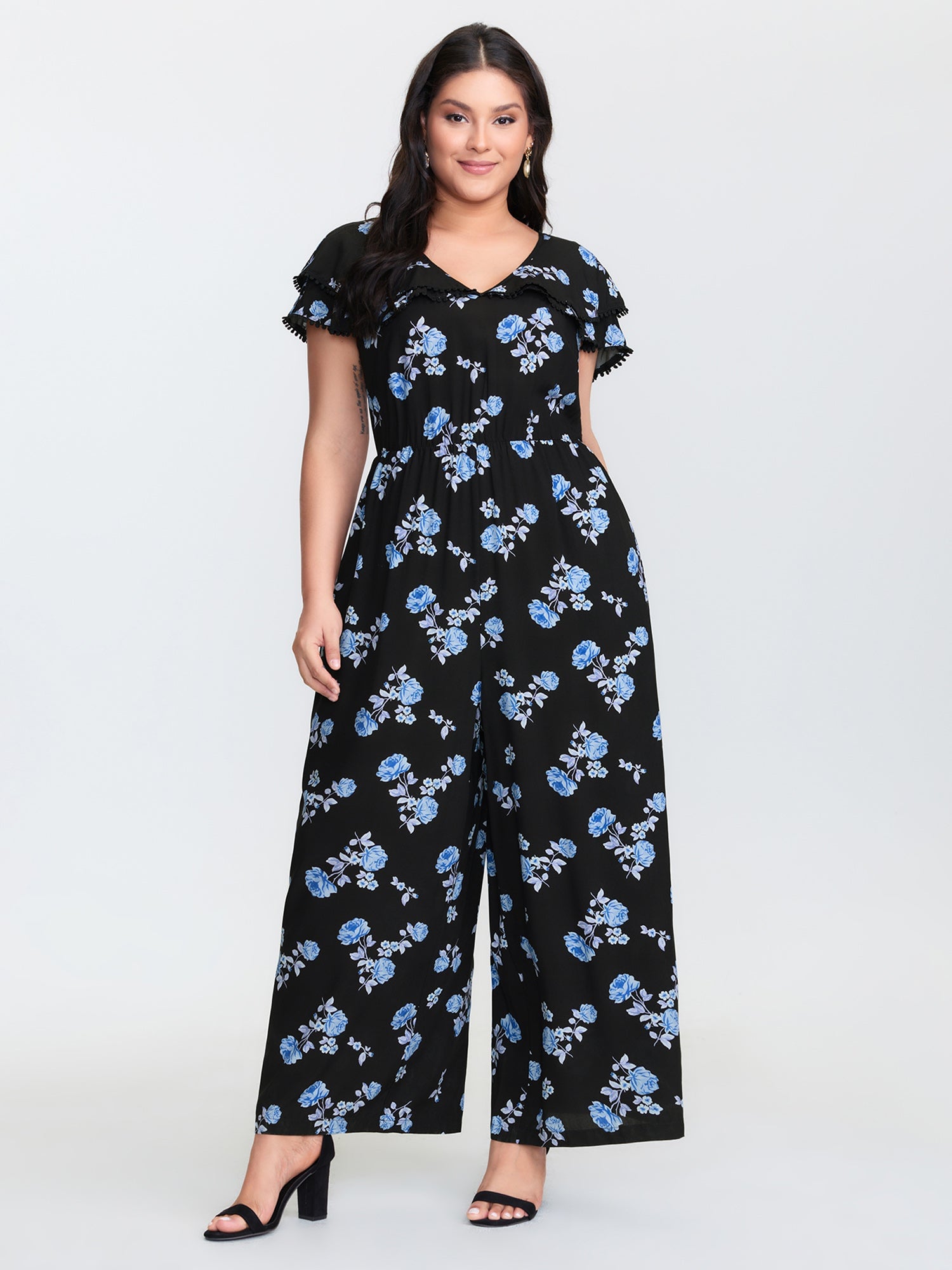Woman in black floral jumpsuit with blue flowers and short ruffled sleeves, standing full length