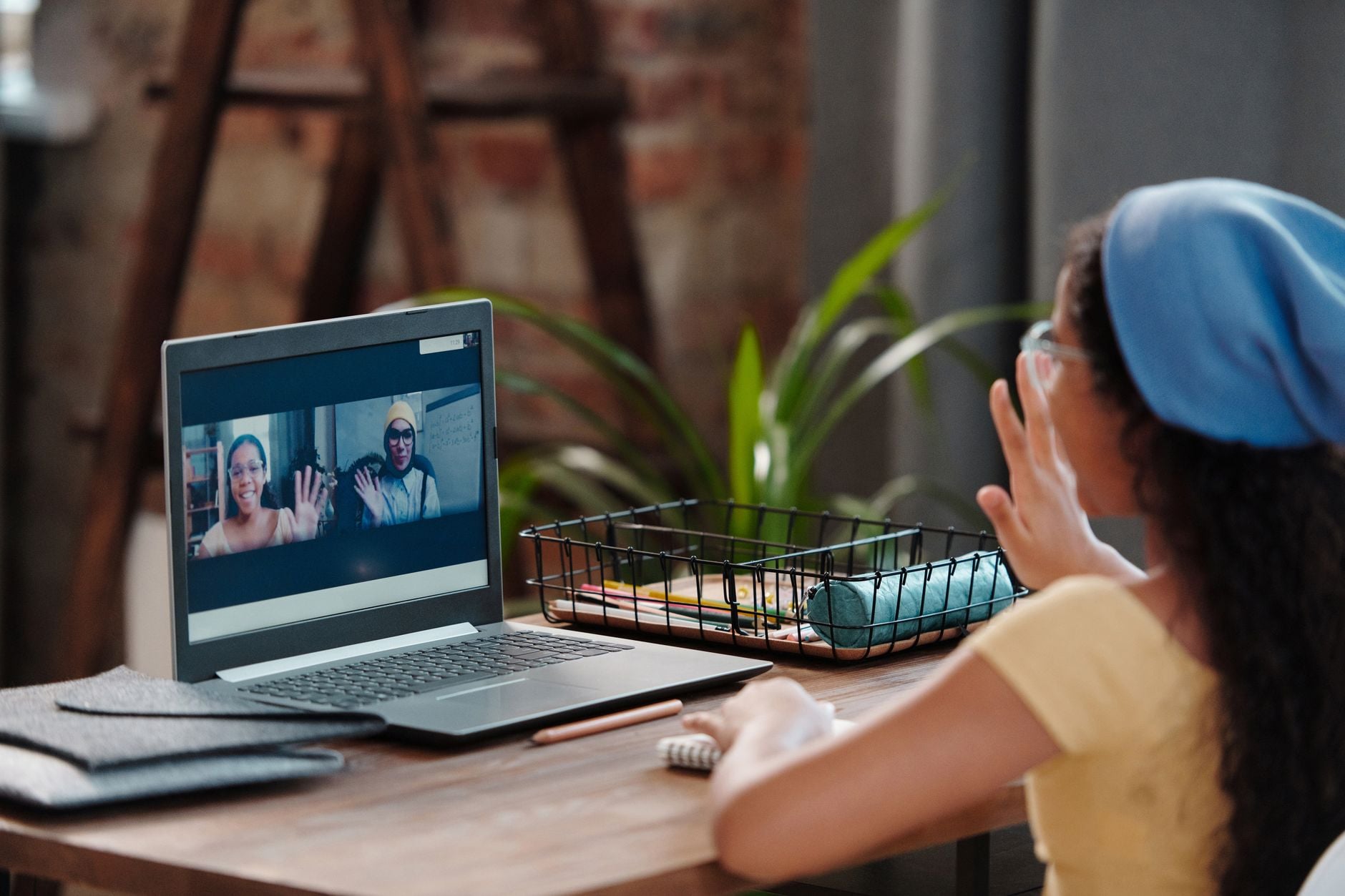 Person in blue cap waving at laptop screen during video call with two people indoors at wooden desk