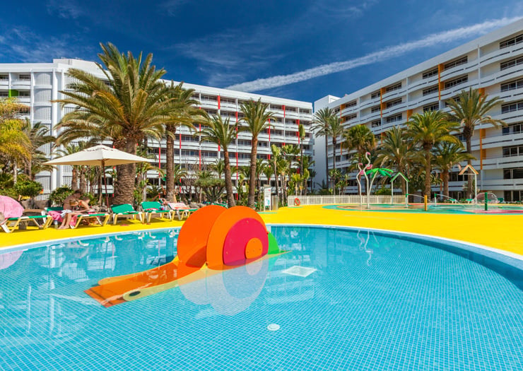 Piscina infantil con tobogán colorido rodeada de palmeras y edificios de hotel de varios pisos