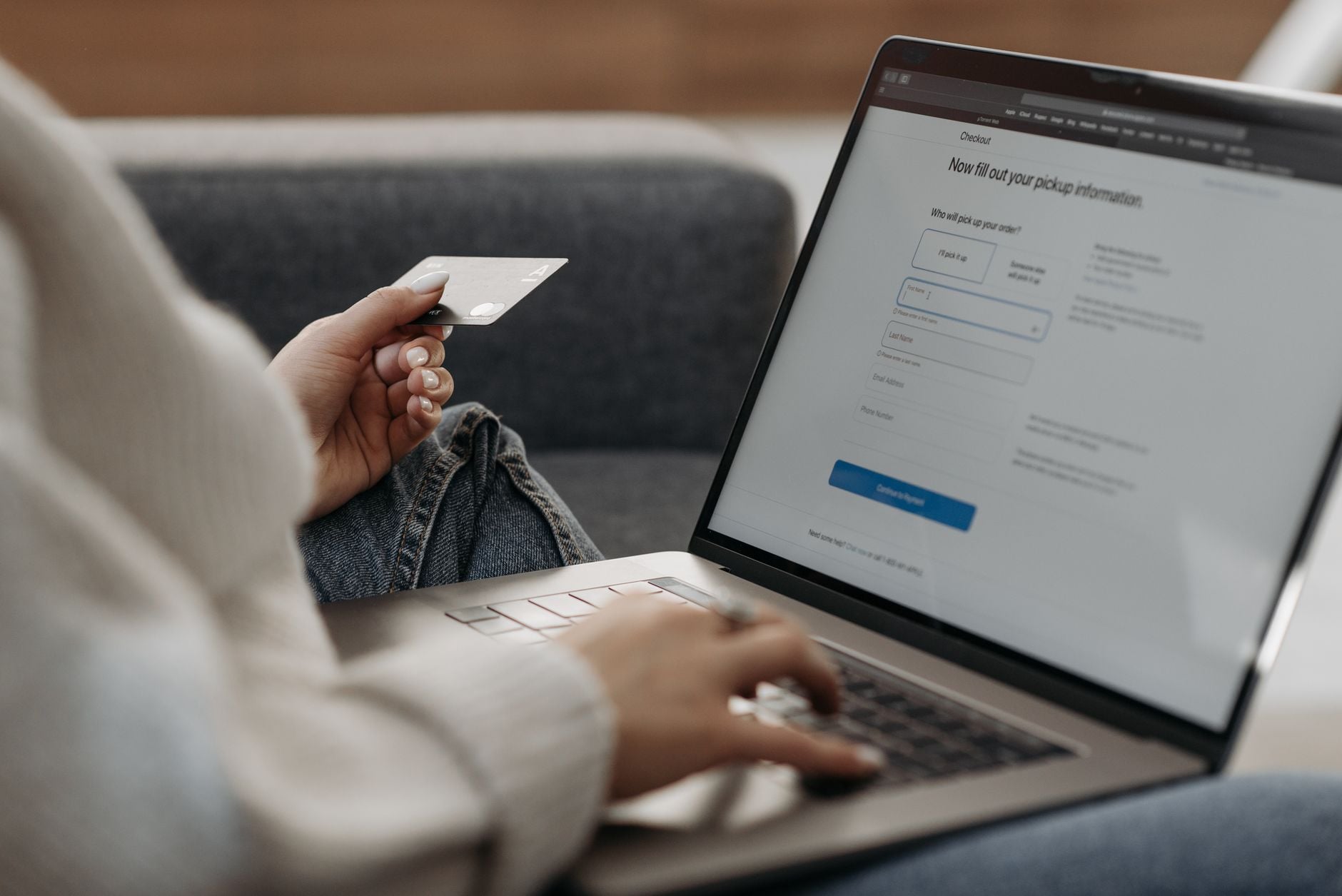 Osoba w białej bluzie siedząca na kanapie trzymająca białą kartę płatniczą przed ekranem laptopa z formularzem checkout
